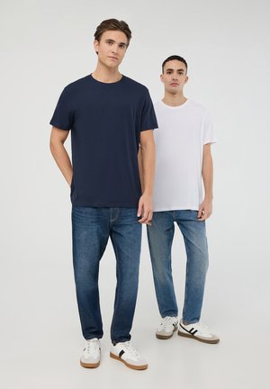 Due uomini stanno fianco a fianco indossando t-shirt basic a girocollo: una blu navy e una bianca, insieme a jeans di denim larghi con scarpe da ginnastica. Design semplice, texture morbida.