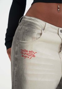 Jeans de mezclilla en gris claro con una textura desgastada, que presentan un logo bordado en rojo de "Don Ed Hardy" en el bolsillo delantero y acentos de metal.