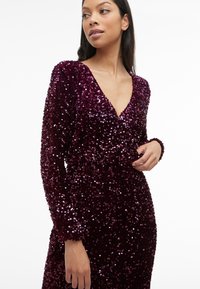 Kvinne i en langermet, dyp burgunderkjole dekket av paljetter med V-hals, poserer mot en ensfarget hvit bakgrunn.