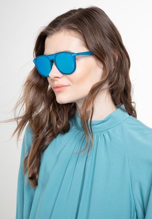 Donna con capelli castani lunghi che indossa occhiali da sole blu brillante e una blusa verde acqua a collo alto, rivolta verso sinistra su sfondo bianco.