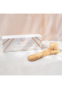 Szczoteczka do twarzy z bambusa z naturalnym włosiem, drewnianym uchwytem oraz białym opakowaniem z złotymi akcentami i geometrycznymi elementami wzorniczymi.