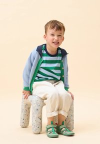 Cardigan azzurro chiaro con dettagli verdi sopra una maglietta a righe bianco e verde. Pantaloni bianchi e scarpe verdi. Seduto su uno sgabello texturizzato e macchiato.
