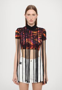Top corto negro con un patrón abstracto colorido. Presenta flecos verticales que se extienden desde el escote. Combinado con pantalones cortos blancos de cintura alta.