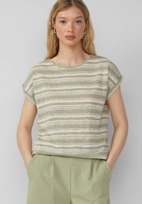 Pasek bluzki z krótkim rękawem w jasnym zielonym i beżowym kolorze, wykonana z fakturowanej tkaniny. Posiada okrągły dekolt i luźny krój, zestawiona z jasn zielonymi spodniami.