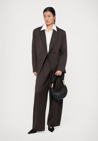 Fato castanho ajustado com pernas largas, camisa branca por baixo e uma mala preta. Apresenta um blazer estruturado e saltos altos pretos pontiagudos.