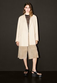 Beige maßgeschneiderter Blazer über einem durchsichtigen schwarzen Top, kombiniert mit beigen Wildleder-Culottes und schwarzen Plateausandalen. Sanfte Texturen, minimalistisches Design.