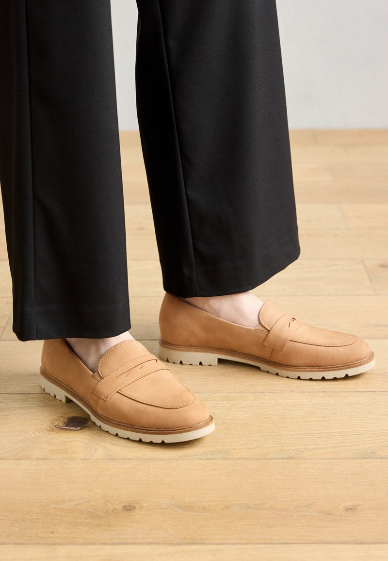 Mocassins en daim beige avec des accents brodés et une semelle en caoutchouc. Associés à un pantalon noir sur un sol en bois.