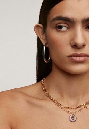 Collares de cadena de oro en capas con un colgante circular y un pendiente en forma de aro metálico liso, sobre un tono de piel neutro.