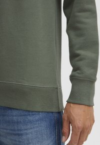 Olive green sweatshirt with a round neck and ribbed cuffs. Features textured fabric and a straight hem. Worn with blue denim jeans.