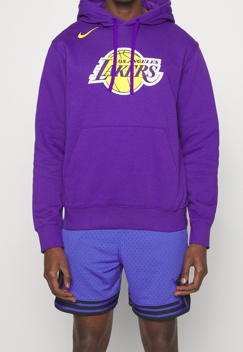 Personne portant un sweat à capuche violet des Los Angeles Lakers avec un logo jaune et un short de basketball violet assorti avec des rayures noires.