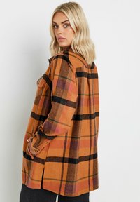 Rutigt skjorta i orange nyanser, med svarta och gröna accenter, knäppta manschetter och en avslappnad passform med sidoklyftor för ökad rörlighet.