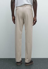 Man wearing beige trousers and black shoes standing on a speckled dark floor against a plain light wall, viewed from the back.