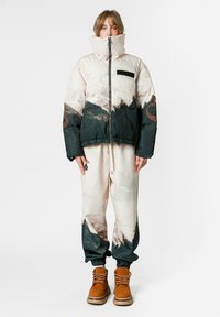 Chaqueta acolchada en beige y verde oscuro con estampado abstracto de montaña; cierre de cremallera y bolsillo en el pecho. Pantalones deportivos crema a juego y botas marrones.