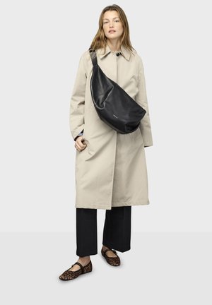 Crna kožna torba za rame s zakrivljenim oblikom, nošena preko bež trench kaputa. Model nosi crne hlače i cipele s leopard printom.
