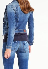 Light blue denim jacket with a fitted design, button closure, and logo patch on the back. Worn with darker blue jeans.