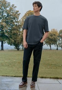 Donkergrijze t-shirt met korte mouwen, zwarte spijkerbroek, bruine schoenen met een gestructureerde zool, staand buiten op een natte stenen ondergrond met bomen op de achtergrond.