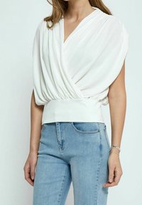 Top blanco con diseño cruzado, escote amplio y cintura fruncida. Combinado con unos jeans ajustados de color azul claro.