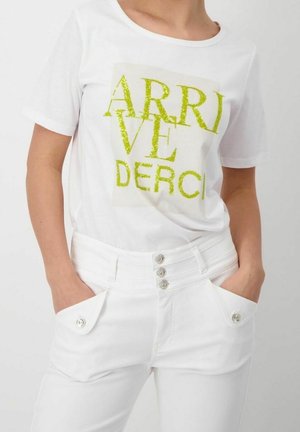 Kobieta w białej koszulce z zielonymi, błyszczącymi literami oraz białych wysokich spodniach z guzikami i kieszeniami, ręce w kieszeniach.