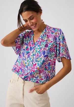 Donna che sorride con la mano dietro al collo indossando una blusa a fiori colorata a maniche corte e pantaloni beige chiaro.