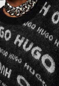 Zwarte trui met witte "HUGO" tekstpatroon, voorzien van een ronde hals en een gestructureerde gebreide stof; aangevuld met een grove zilveren kettingkraag.