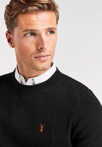Ung man med kort blont hår som bär en svart stickad tröja över en vit skjorta med krage, som tittar något åt vänster mot en enkel bakgrund.