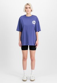 Fioletowy, oversize'owy t-shirt z białym napisem "ALPHA INDUSTRIES", krótki rękaw, zestawiony z czarnymi szortami rowerowymi i białymi sneakersami. Białe tło.