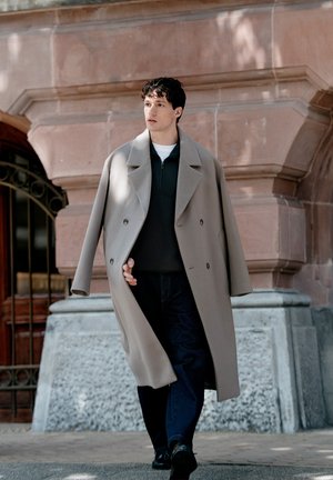 Un homme passe devant un bâtiment en pierre, vêtu d’un long manteau gris, d’un pull foncé, d’un jean et de chaussures noires.