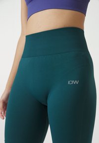 Leggings a vita alta color turchese realizzati in tessuto elasticizzato, con cintura a coste e un piccolo logo grigio sul lato sinistro.