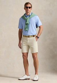 Polo azul claro, pantalones cortos beige, suéter verde sobre los hombros, cinturón marrón y beige, zapatos de vestir blancos, gafas de sol oscuras.