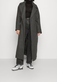 Lang, zwart-wit geblokte jas met brede kraag, dubbelrijge knopen en een losse, oversized pasvorm. Gedragen over een aangesloten zwart shirt en spijkerbroek.