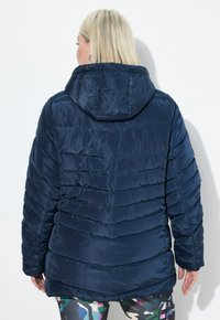 Chaqueta de plumas azul marino con capucha, textura acolchada y un ajuste holgado. Presenta mangas largas y un dobladillo redondeado.