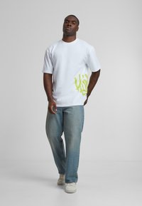 White cotton t-shirt featuring a green abstract design on the left side, paired with light blue denim pants and white sneakers.