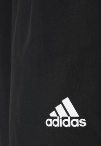 Mustat kangas urheiluvaatteet, joissa on näkyvä valkoinen Adidas-logo ja kolme diagonaalista raitaa. Sujuva tekstuuri ja virtaviivainen muotoilu.