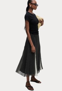 Un haut noir à manches courtes associé à une jupe midi à pois noirs et blancs, avec un design fluide et une texture transparente, accessoirisé d'une pochette en osier.