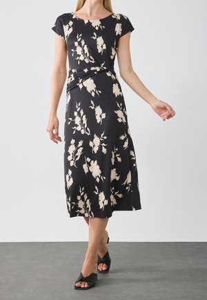 Kvinde iført en sort midi-kjole med beige blomsterprint og korte ærmer, kombineret med sorte åbne hæl-sandaler, stående på en grå gulv.