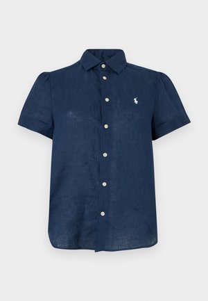 Γυναίκα που φοράει μπλε σκούρο πουκάμισο με κοντά μανίκια και κουμπιά, λευκά σορτς, λευκά σανδάλια, κρατώντας μια πράσινη και κίτρινη υφαντή τσάντα.