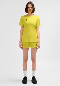 Żółty sportowy komplet koszulka i spodenki wykonane z lekkiego materiału, z logo na klatce piersiowej i udzie. Noszony z czarnymi trampkami i białymi skarpetkami.