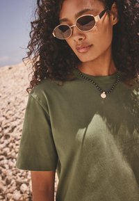 T-shirt en coton vert olive présentant une coupe décontractée, un col rond et des manches courtes. Accessoirisé d'un collier de perles noires et de lunettes de soleil rondes.