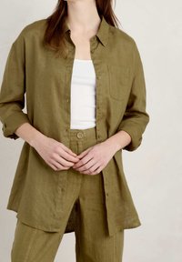Olive green linen shirt with long sleeves, button-down front, and a chest pocket, styled with matching high-waisted pants. Smooth texture.