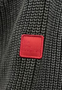 Adīta tumši pelēka džempera auduma ar strukturētu rakstu. Tajā ir sarkans kvadrātveida ielāps ar reljefā iespiestu tekstu "the new originals."