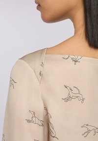Top beige con una vestibilità comoda, caratterizzato da un sottile motivo di contorni grigi di cani in corsa. Tessuto morbido con scollatura ampia e maniche trasparenti.