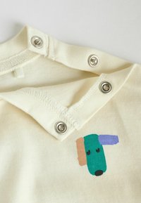 Tissu crème clair avec des boutons-pression métalliques ; présente un graphisme d'un chien stylisé en bleu sarcelle, marron et violet près du col.