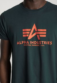Mørkegrøn t-shirt lavet af bomuld med et orange grafisk logo, der viser "ALPHA INDUSTRIES" og trekantede designdetaljer på fronten.