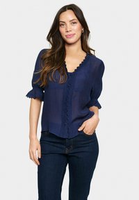 Blusa blu navale con scollo scallop e tessuto testurizzato, con chiusura a bottoni sul davanti e maniche arricciate. Abbinata a jeans scuri.