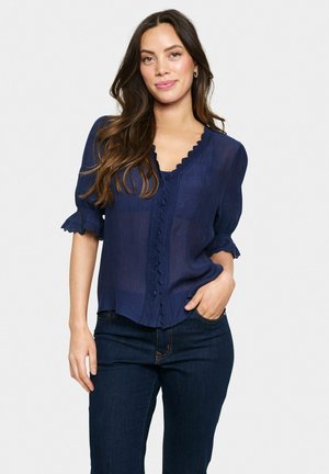Marineblaue Bluse mit gewelltem Ausschnitt und strukturiertem Stoff, mit Knopfverschluss vorne und Rüschensäumen. Kombiniert mit dunklen Jeans.