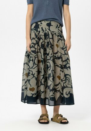 Frau trägt einen gestuften, gemusterten Midi-Rock in Beige und Schwarz mit braunen Akzenten, kombiniert mit sandfarbenen Sandalen mit Schnallen und einem blauen Kurzarmoberteil.