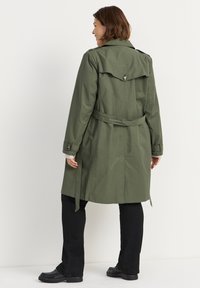Olijfgroen trenchcoat met een achterflap en riem; heeft knoopdetails en lange mouwen, gecombineerd met zwarte broek en schoenen.