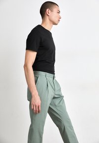 T-shirt noir ajusté associé à un pantalon taille haute plissé de couleur vert clair. Modèle présenté de profil sur un fond neutre.