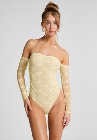 Off-shoulder bodysuit in doorschijnend gele kant, met lange mouwen en een bloemenpatroon, en een hoge beenuitsnijding.