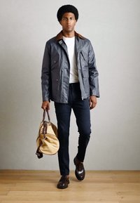 Veste gris charbon avec col marron, chemise blanche, pantalon bleu marine et mocassins marron. Modèle tient un sac beige, debout sur un sol en bois.